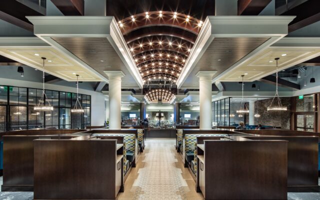 A restaurant interior featuring a central aisle flanked by dark wood booths with patterned cushions, leading to a service area under a vaulted, lit ceiling.