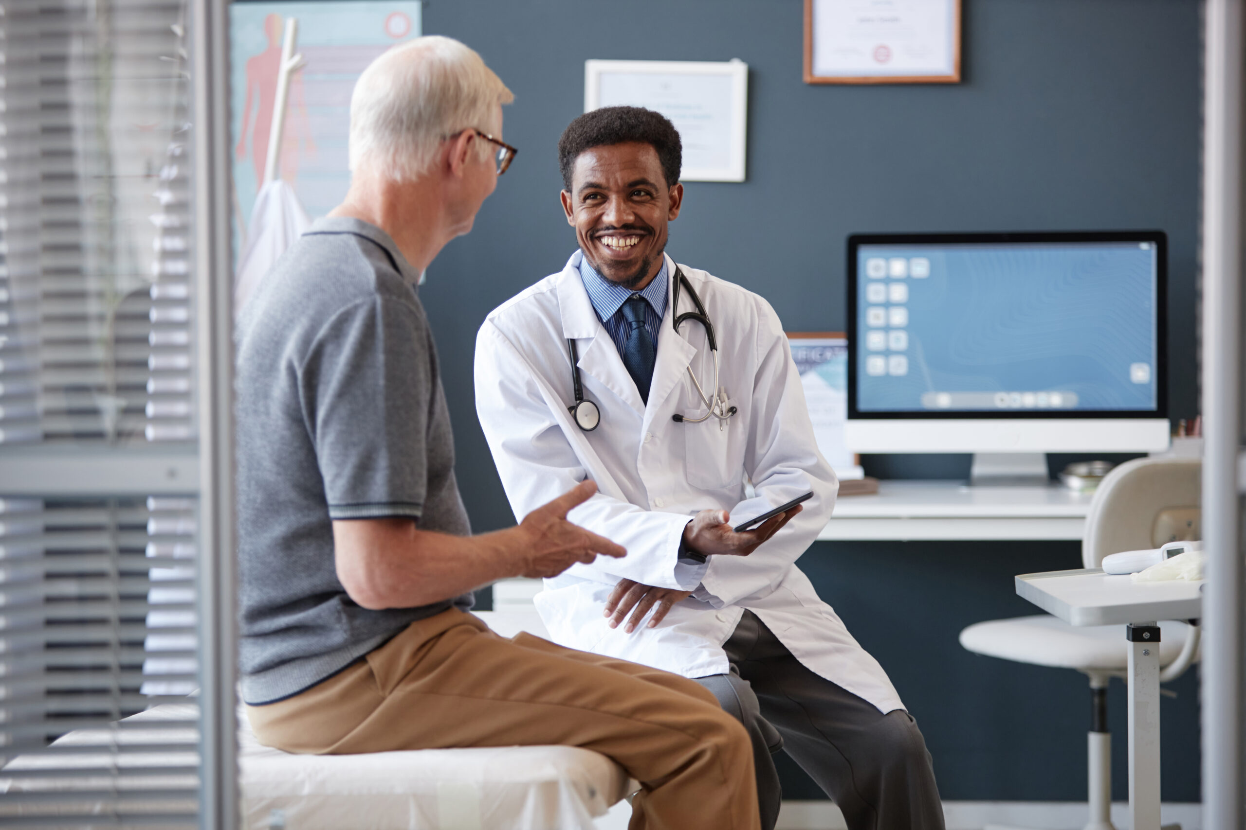 Smiling Doctor Holding Tablet Talking to Patient in Consultation